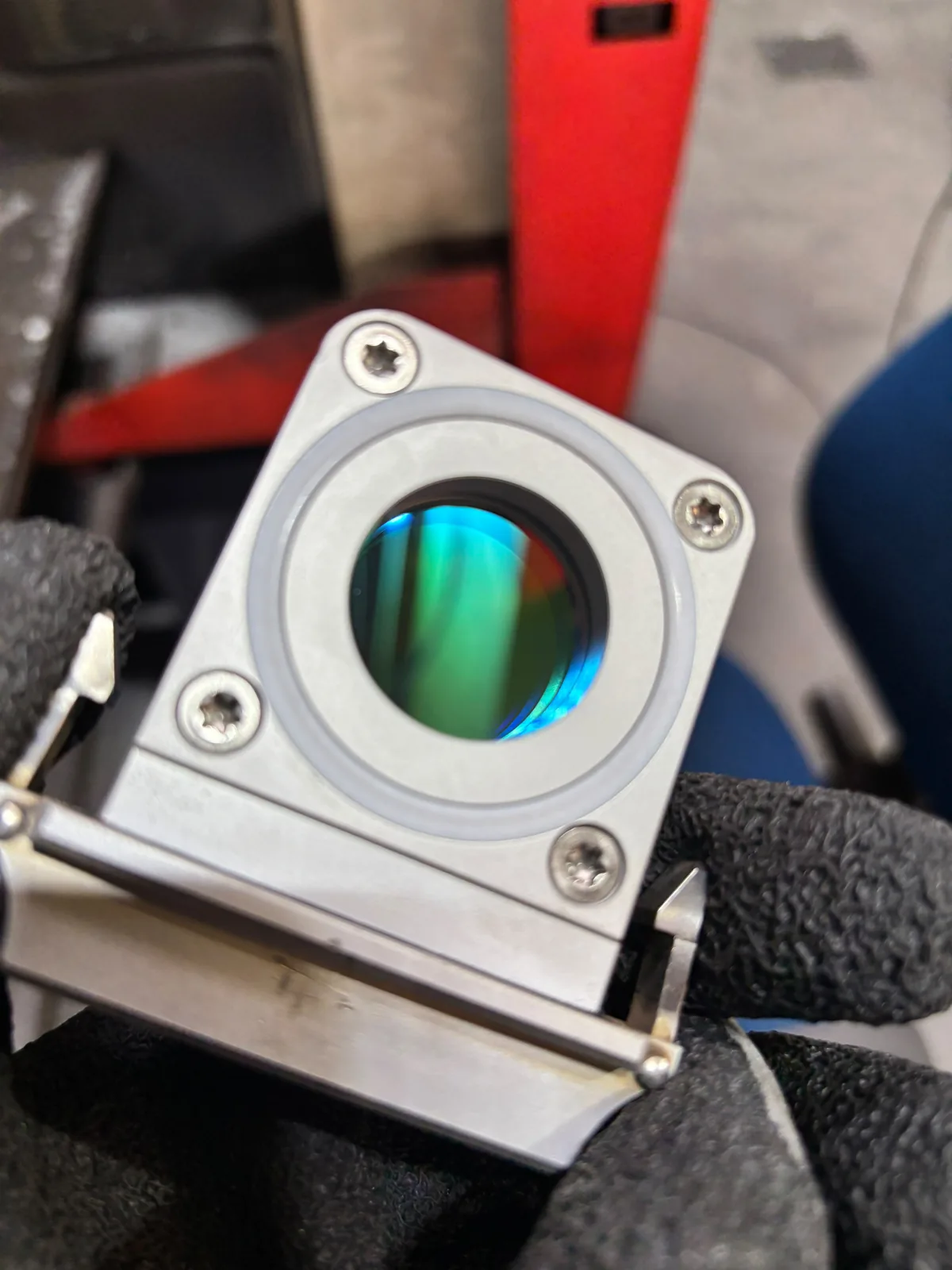 Rechteckige Optikkassette in einer behandschuhten Hand, die Beschichtung schillert grün
